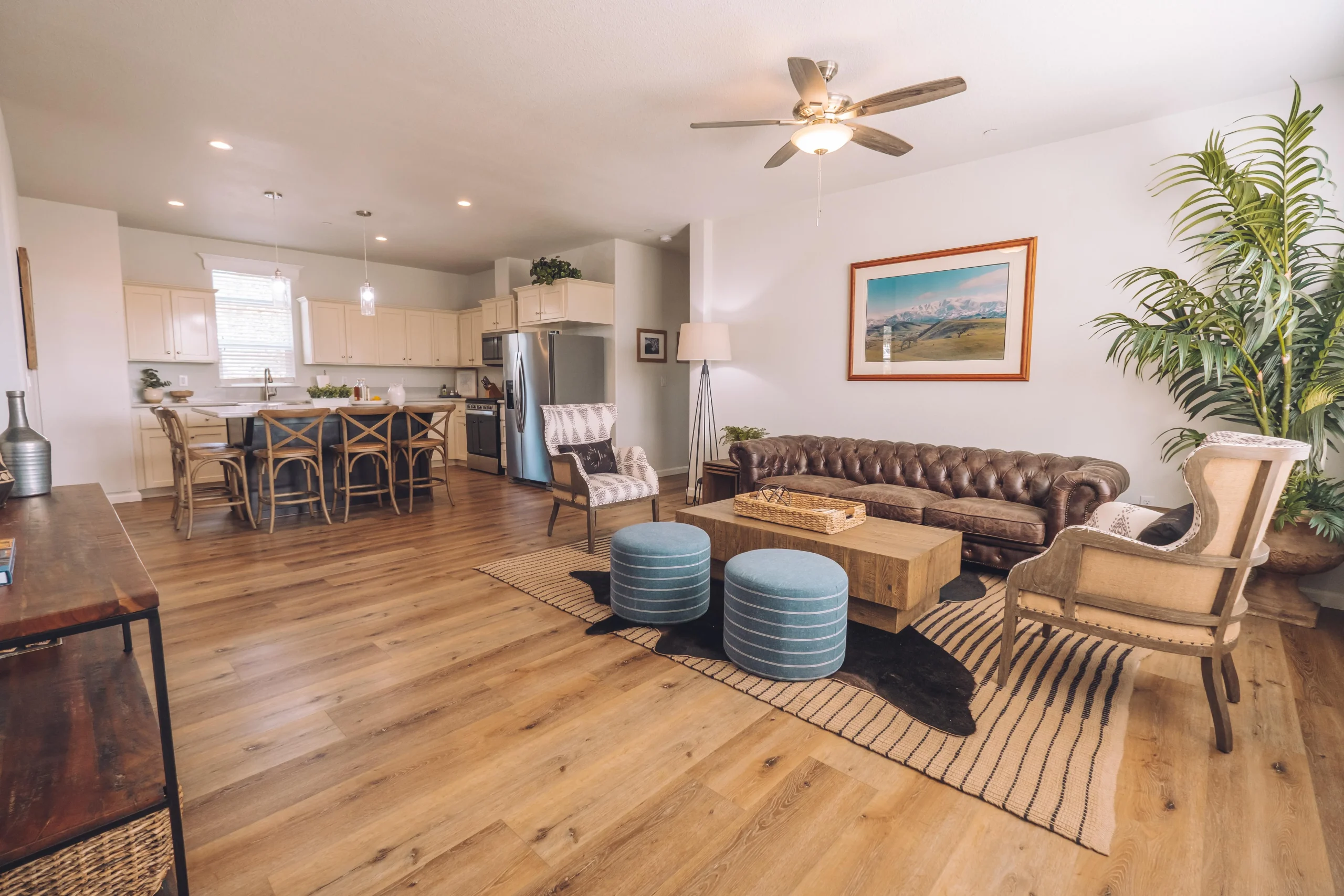Open-plan living room and kitchen featuring a brown leather sofa, upholstered armchairs, a wooden coffee table, blue ottomans, hardwood flooring, and cream kitchen cabinetry.